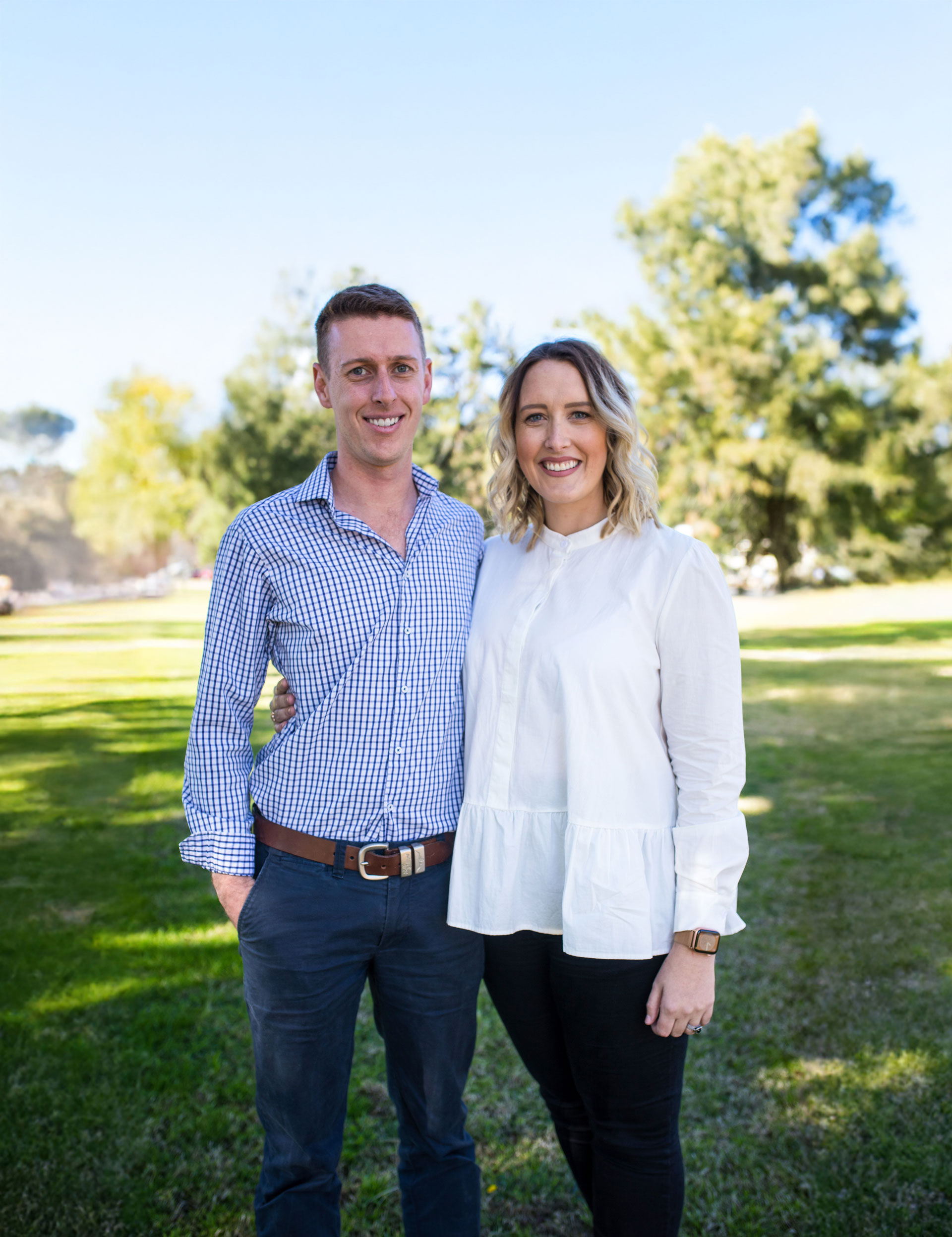 Smiling man and woman standing outdoors with trees in the background.