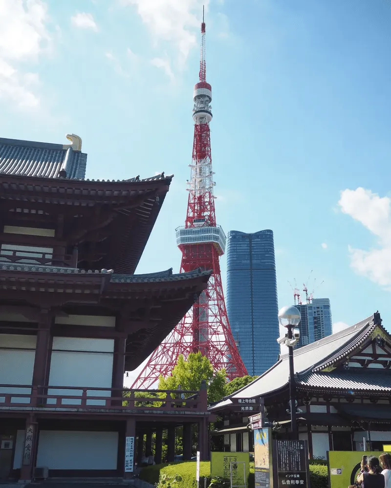 The Tokyo Tower against a blue sky, framed by traditional Japanese buildings in the foreground. Modern skyscrapers are visible in the background.