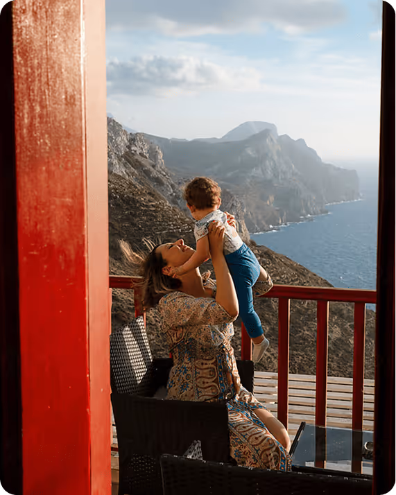 Woman joyfully lifts a child on a sunlit balcony overlooking a scenic coastal cliff and ocean under a blue sky with clouds.