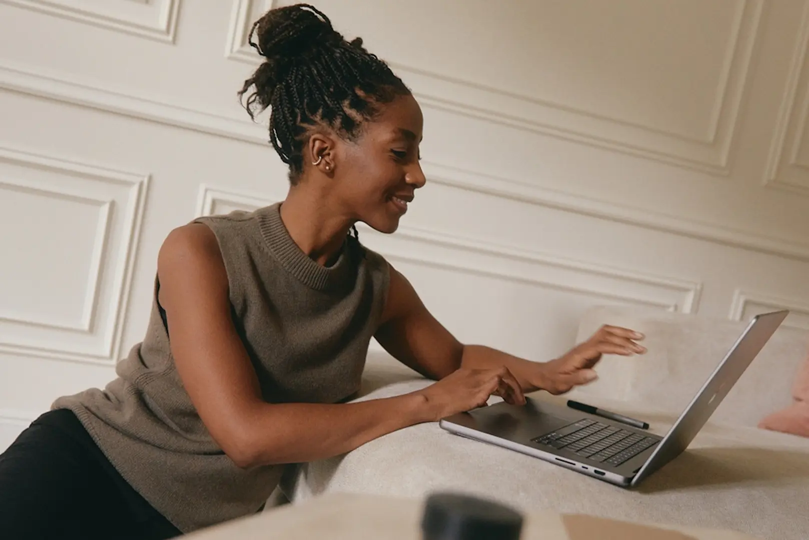 A smiling woman with braided hair sits on a sofa, typing on a laptop. The room has elegant white paneling, creating a cozy and professional atmosphere.