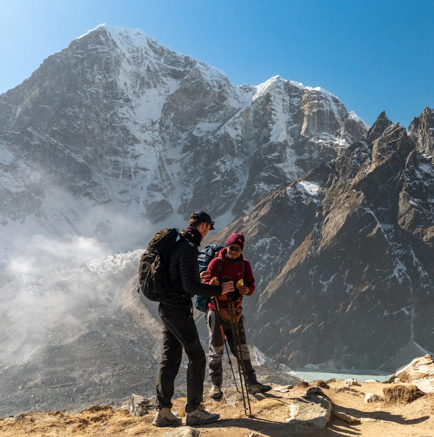 Two hikers stand on a rocky trail in front of towering snow-capped mountains. The sky is clear, conveying a sense of adventure and awe.
