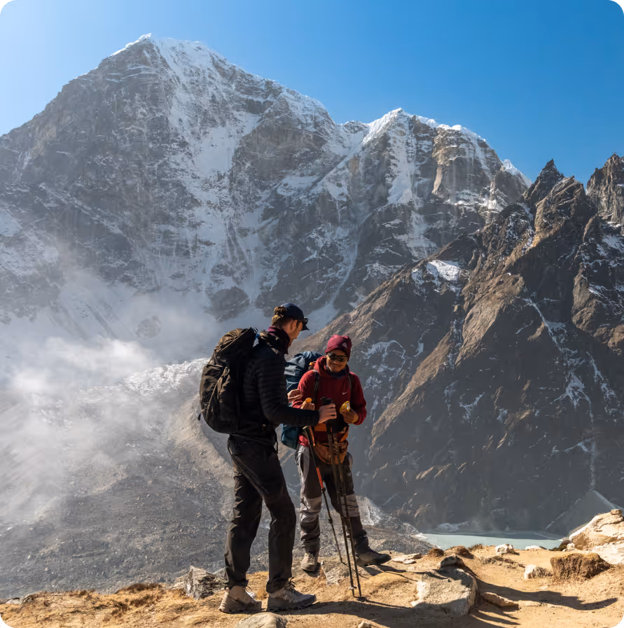 Two hikers stand on a rocky trail in front of towering snow-capped mountains. The sky is clear, conveying a sense of adventure and awe.