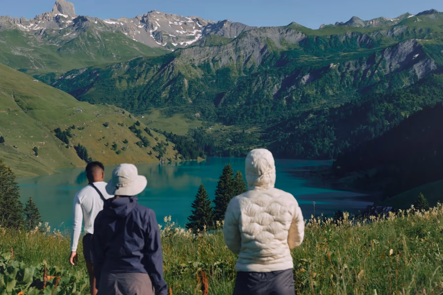 Three people in outdoor gear face a turquoise lake surrounded by lush green hills and distant snow-capped mountains under a clear blue sky.