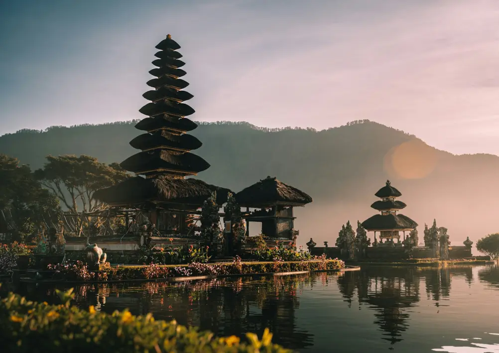 Pagoda-style temple with tiered roof by a reflective lake at sunrise. Misty hills in the background, creating a tranquil and serene atmosphere.