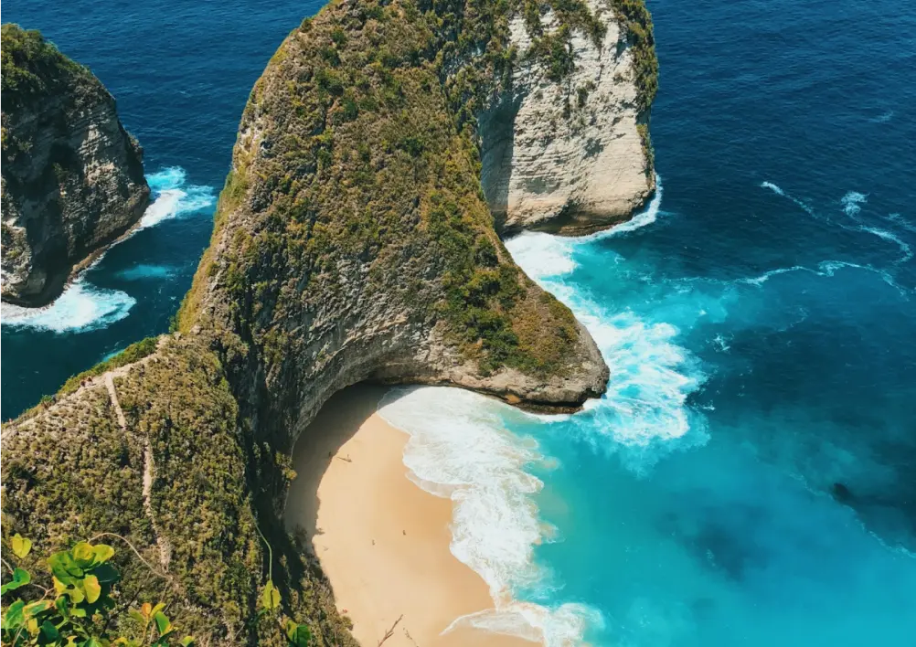 Scenic beach with turquoise waters, dramatic cliffs, and lush green rock formations. A serene sandy shore lined with palm trees under a clear sky.
