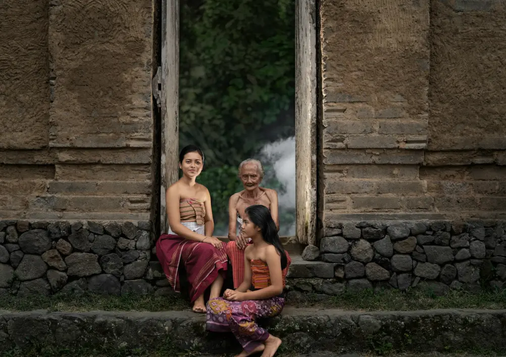 Three people in traditional attire sit on stone steps of an ancient building, conveying a sense of heritage and tranquility. Lush greenery in the background.