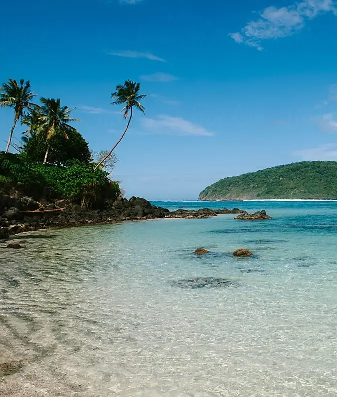 Tropical beach with clear turquoise water, palm trees, and rocky shoreline. A lush green hill rises in the distance under a bright blue sky. Tranquil scene.