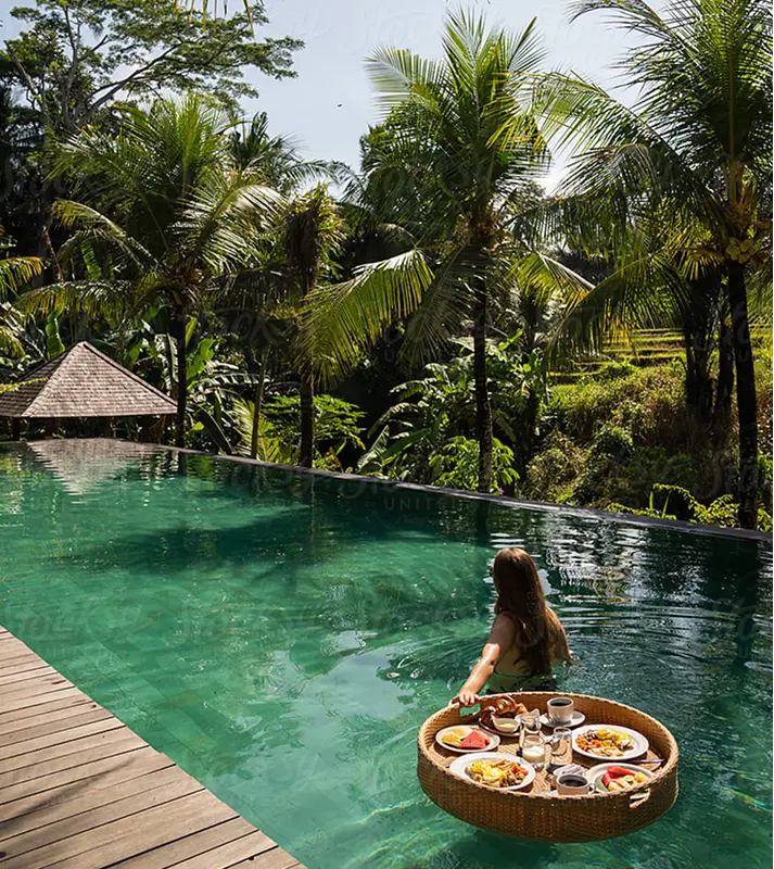 A woman in a pool pushes a round wicker tray with breakfast. Palm trees surround the serene, tropical setting, under a sunny blue sky.