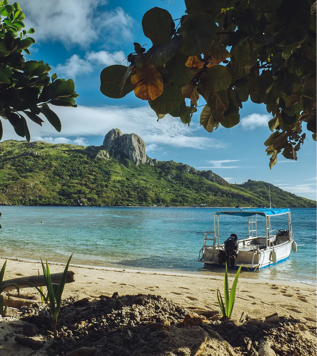 Serene Fiji beach scene with a motorboat anchored on clear, turquoise water. Lush green island and rocky hill in the background, framed by leafy branches.