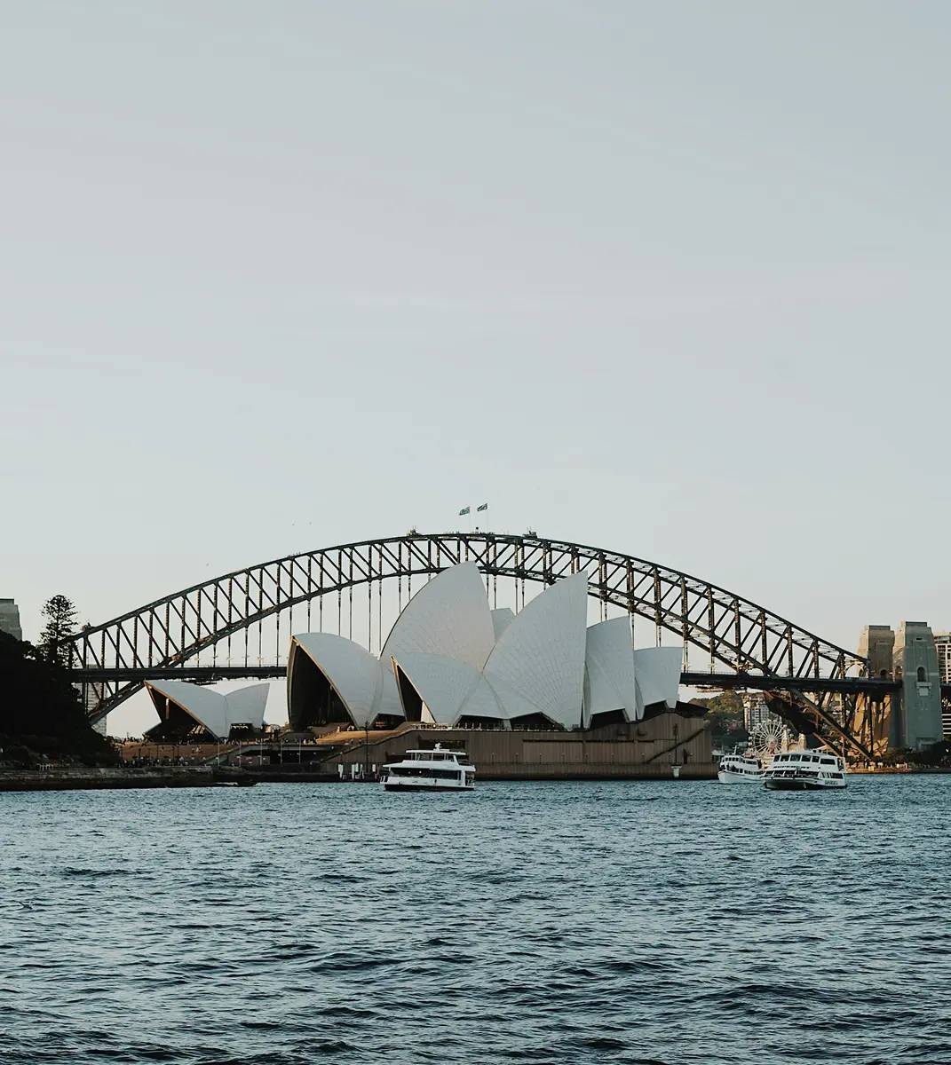 Sydney Opera House and Harbour Bridge at sunset, viewed from the water. Boats are in the foreground, and the sky is clear and softly lit.
