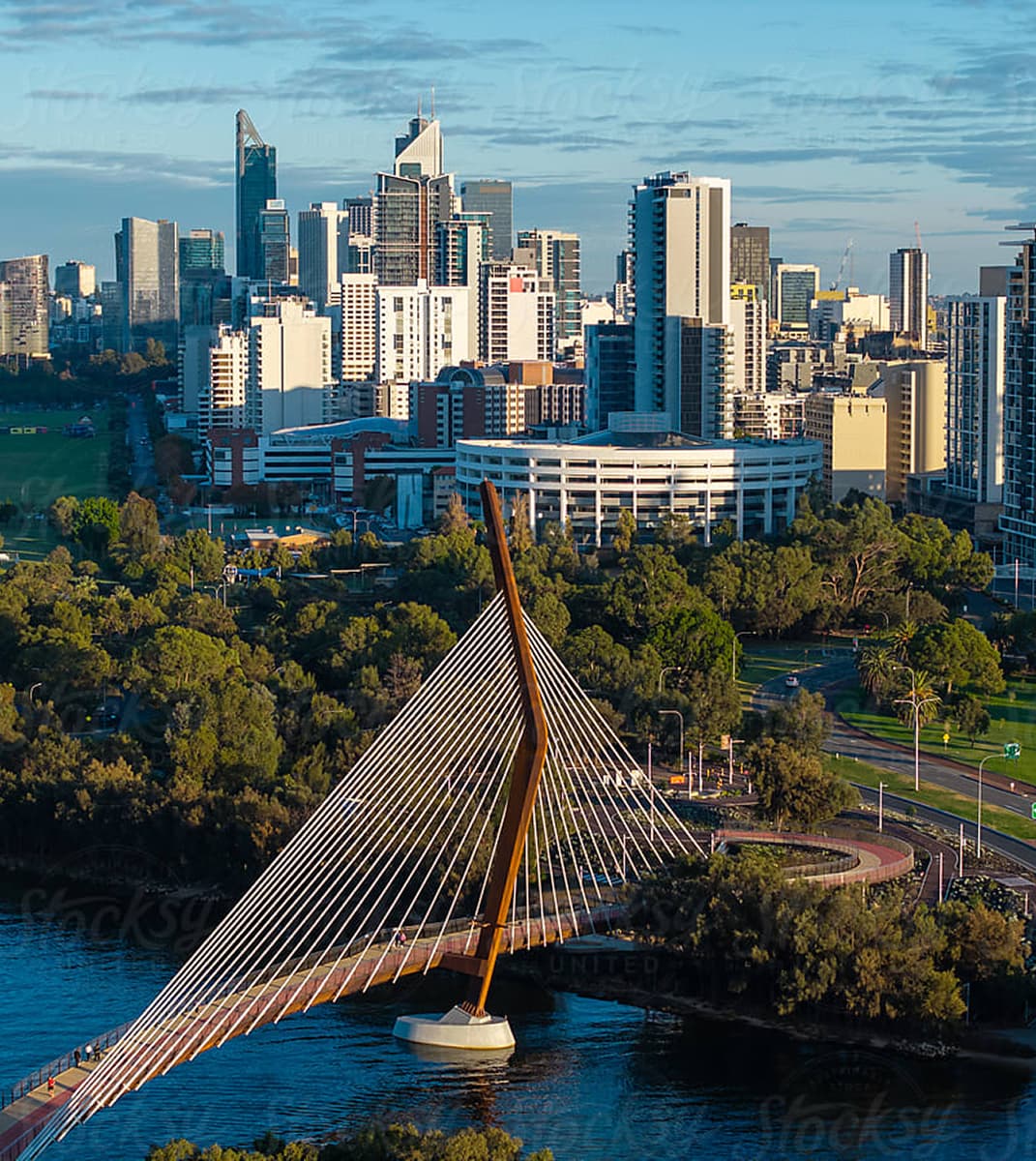 The Boorloo bridge spans across a river with a backdrop of a bustling cityscape. Tall buildings and lush greenery are bathed in soft, golden sunlight.