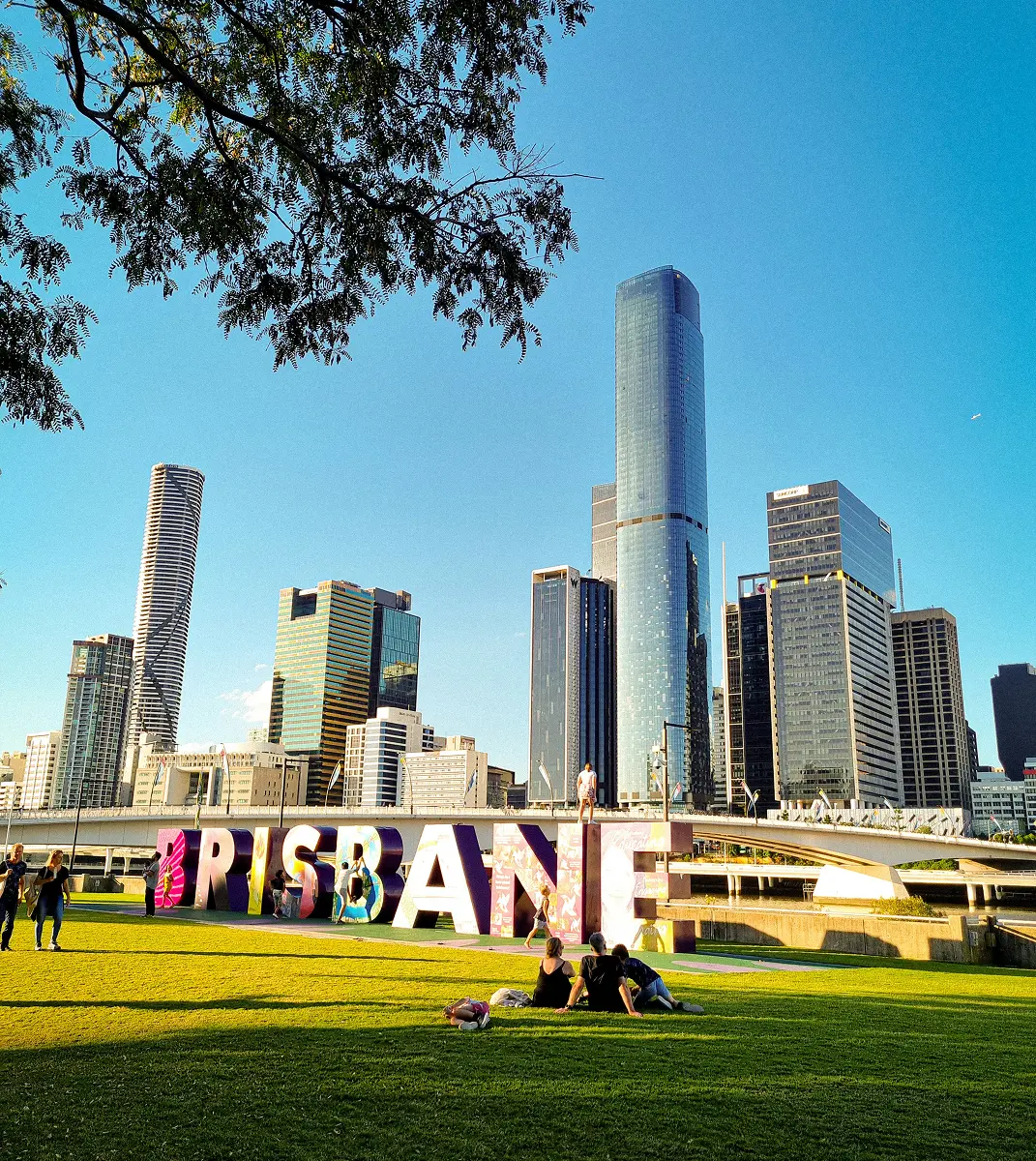Skyline of Brisbane with modern skyscrapers under a clear blue sky. Giant colorful letters spelling "BRISBANE" in a park, people relaxing on grass.