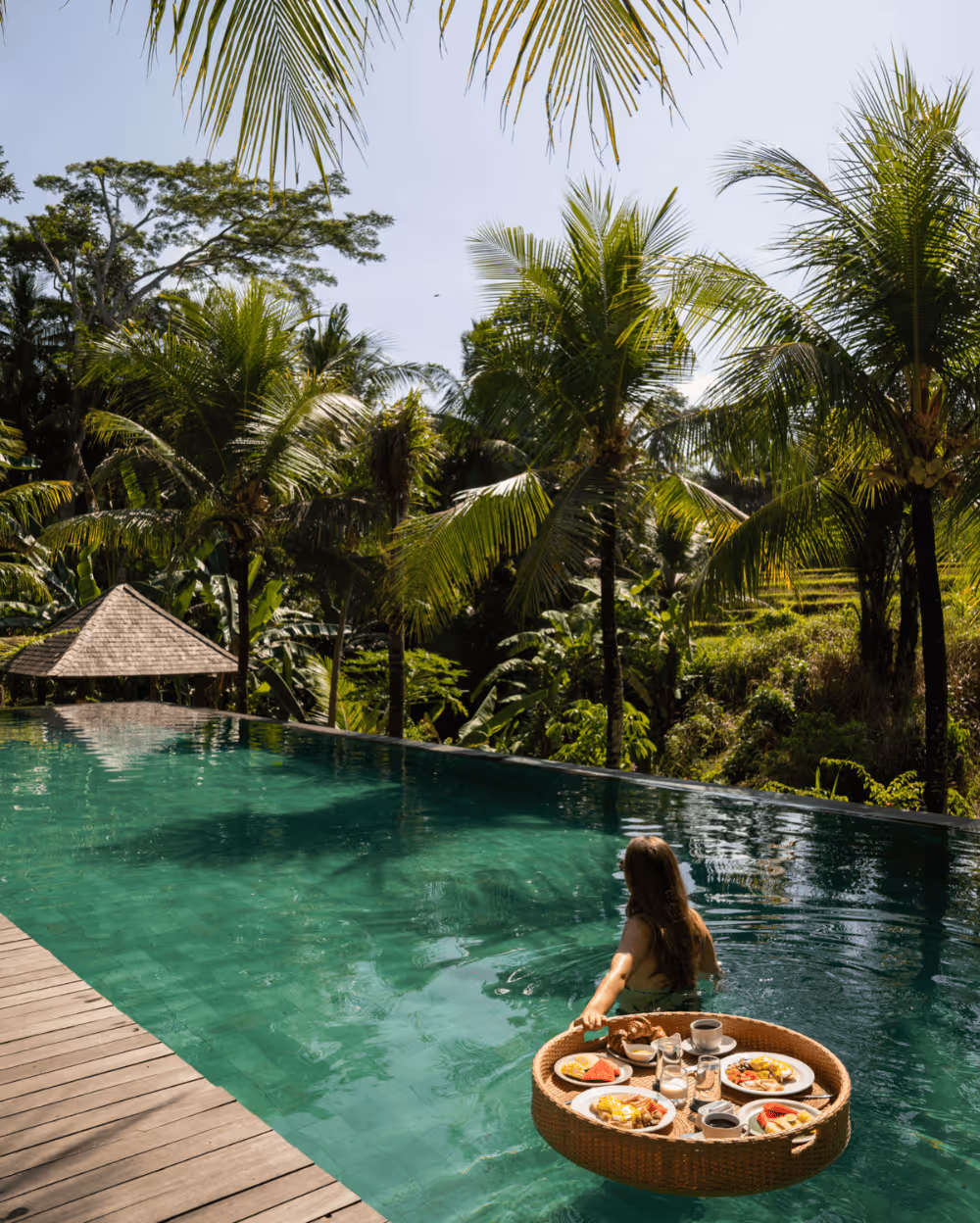 A woman in a pool pushes a round wicker tray with breakfast. Palm trees surround the serene, tropical setting, under a sunny blue sky.