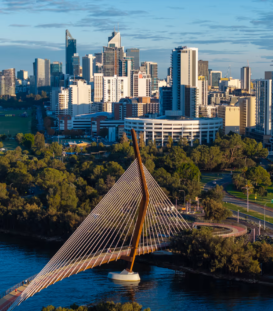 The Boorloo bridge spans across a river with a backdrop of a bustling cityscape. Tall buildings and lush greenery are bathed in soft, golden sunlight.