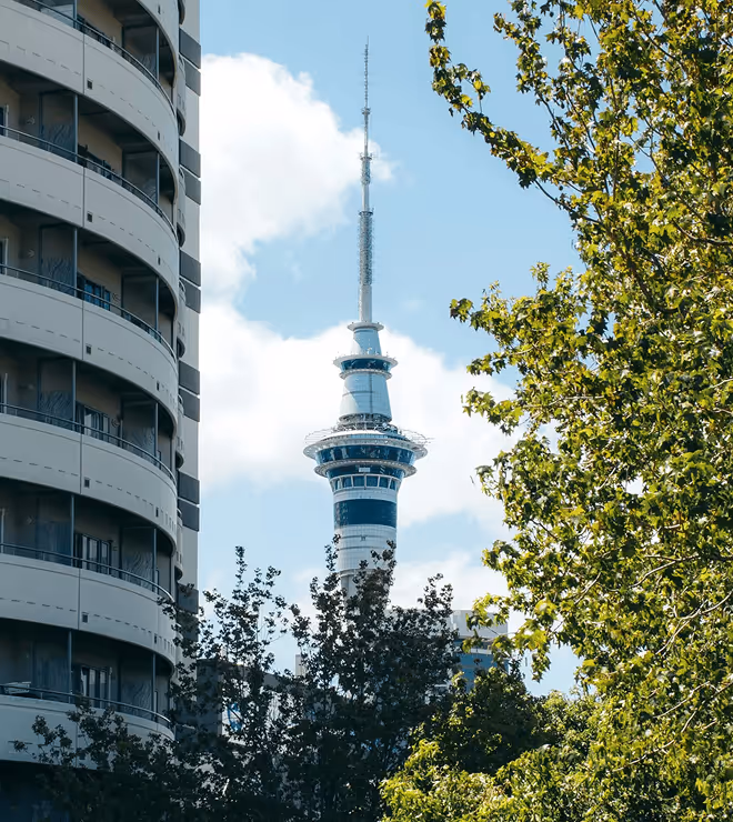 Sky tower against a blue sky with clouds, framed by modern curved building on the left and leafy trees on the right. Urban scene.
