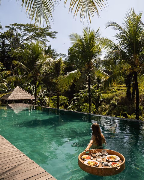 A woman in a pool pushes a round wicker tray with breakfast in Bali. Palm trees surround the serene, tropical setting, under a sunny blue sky.