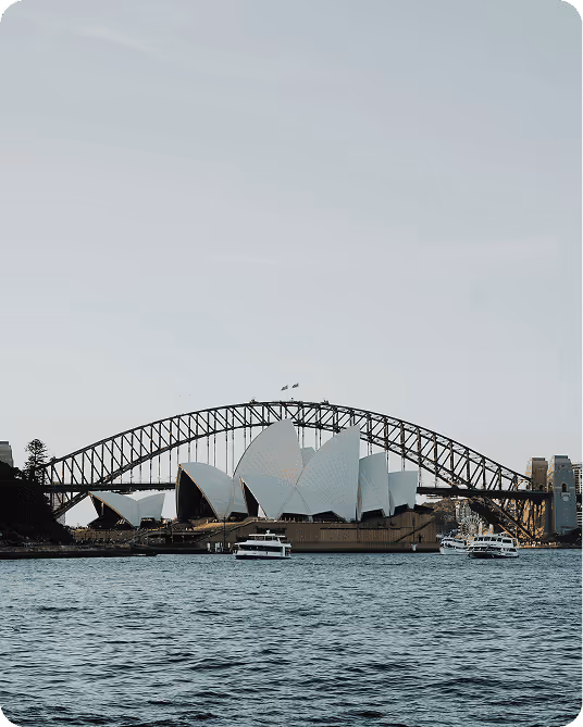 Sydney Opera House and Harbour Bridge at sunset, viewed from the water. Boats are in the foreground, and the sky is clear and softly lit.