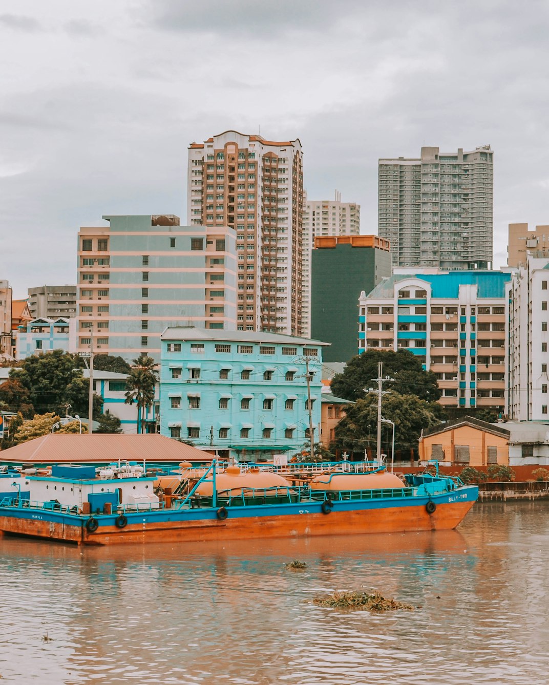 A blue and brown boat on the Pasig River near Fort Santiago in Intramuros, with Manila city buildings behind.