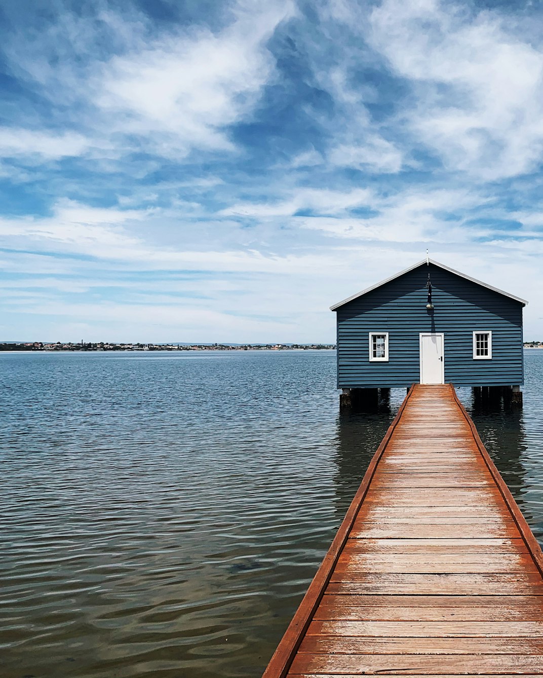 A blue boatshed at the end of a long wooden jetty on the Swan River in Perth under a bright sky.