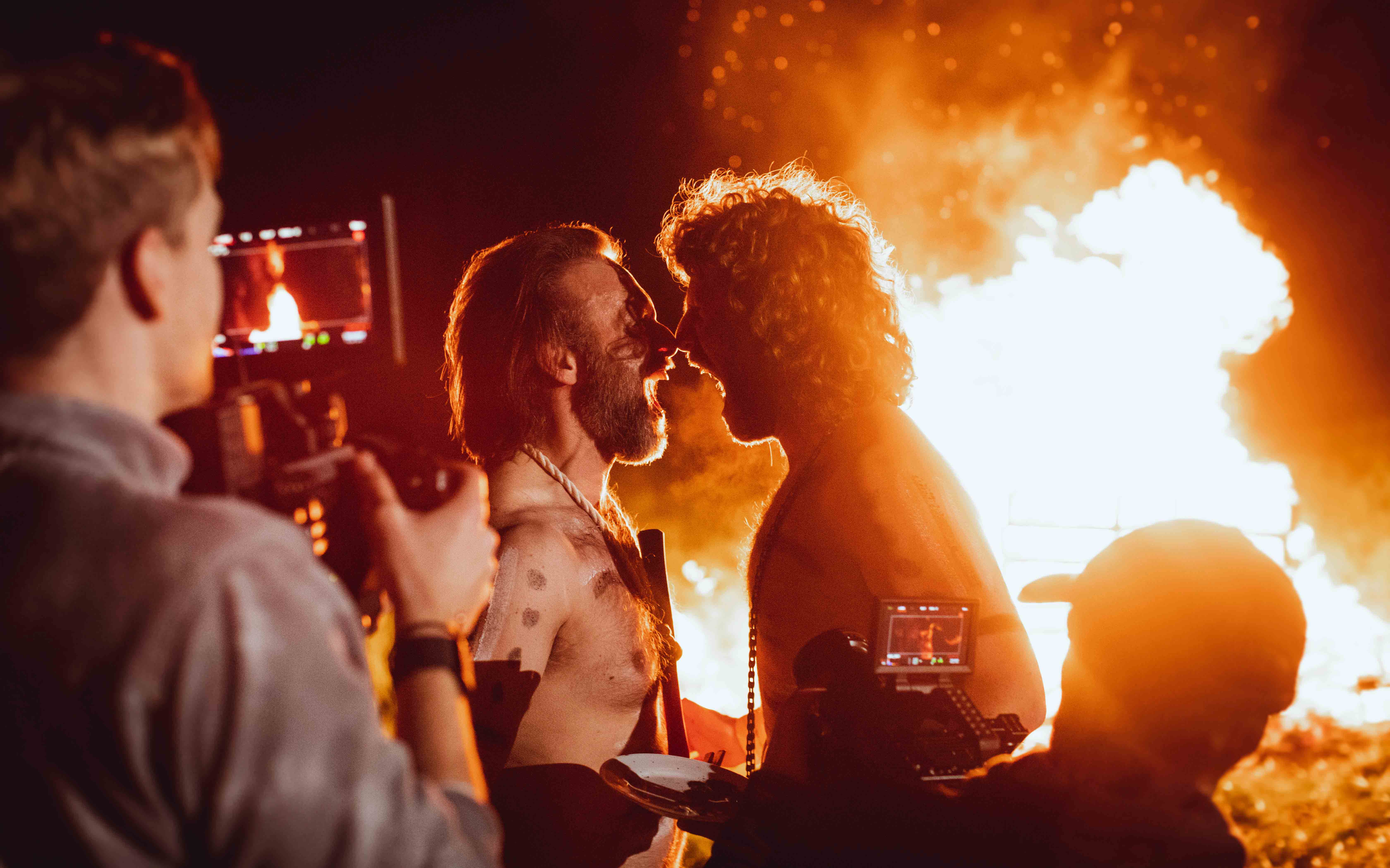 Two men yell at each other in front of a large fire for a comedic video advert. A cameraman can be seen operating a RED cinema camera.
