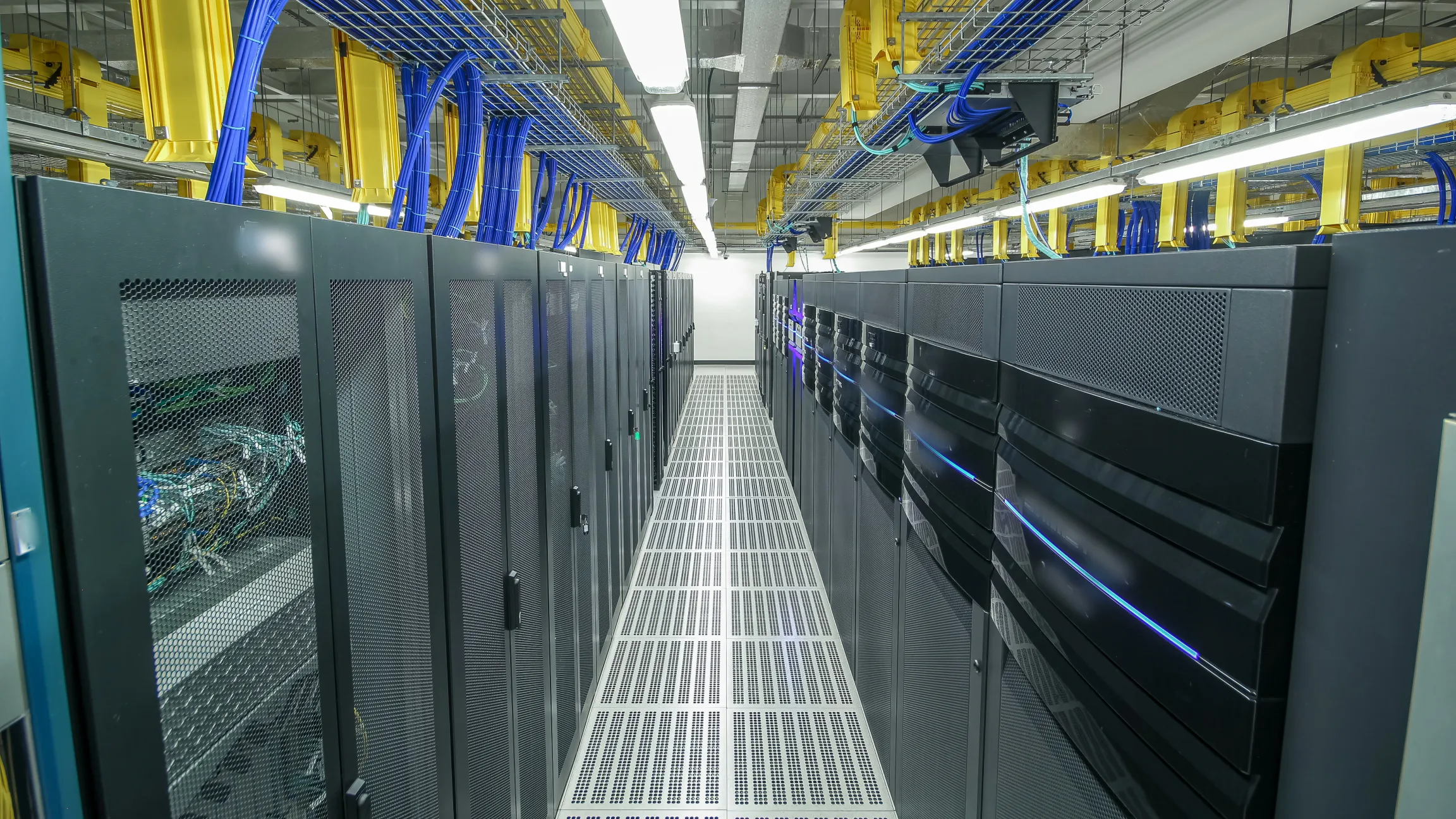 Aisle of server racks in a data center with organized blue and yellow structured cabling overhead.