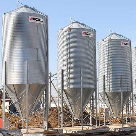 Three tall silver Roxell feed bins or silos with conical bottoms on a construction site under a clear sky.