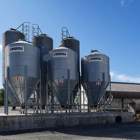 Several large silver Roxell feed bins or silos on a concrete platform under a blue sky.