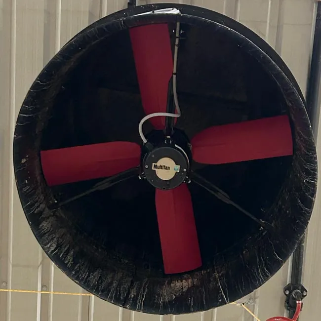 Large industrial ceiling fan with four red blades and a black frame mounted on a corrugated metal roof.