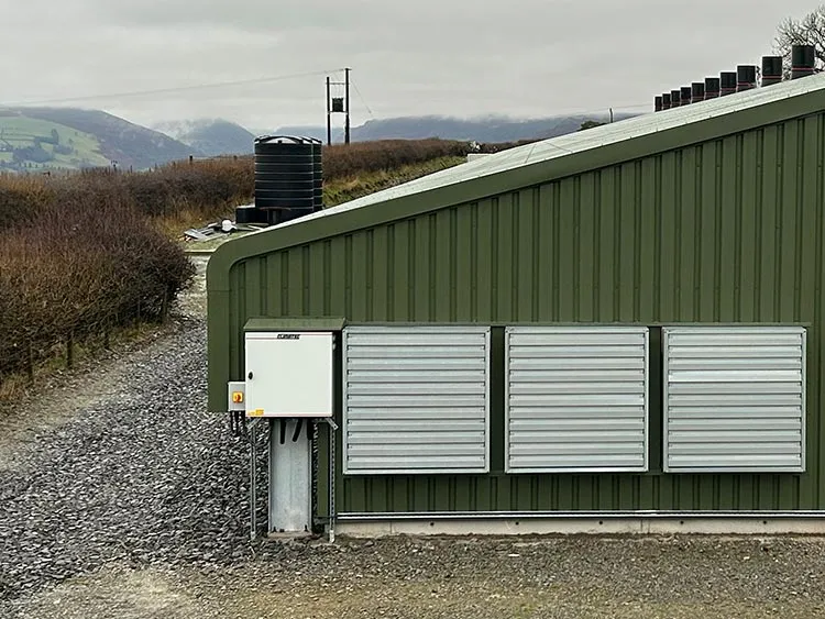 Green industrial building with three metal ventilation panels on its side and gravel pathway nearby.