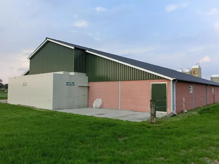 Green and brick agricultural building with adjoining beige equipment housing on a grassy field under a partly cloudy sky.
