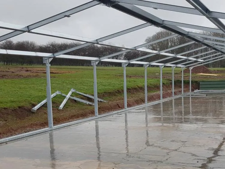 Metal frame structure on a wet concrete foundation in a grassy outdoor area with cloudy sky.