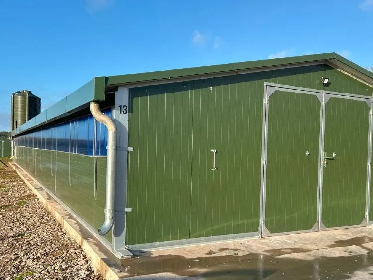 Long green metal agricultural building with large double doors and a nearby silo under a blue sky.