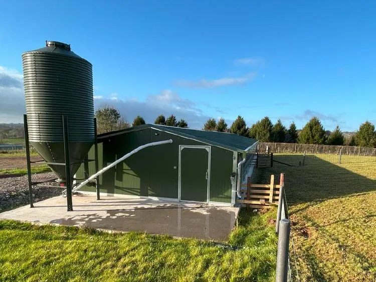 Green metal agricultural shed with attached feed silo and a fenced grassy field under a blue sky.