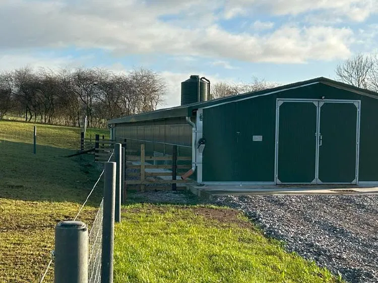 Green agricultural barn with double doors, water tank on the roof, and fenced grassy area under a cloudy sky.
