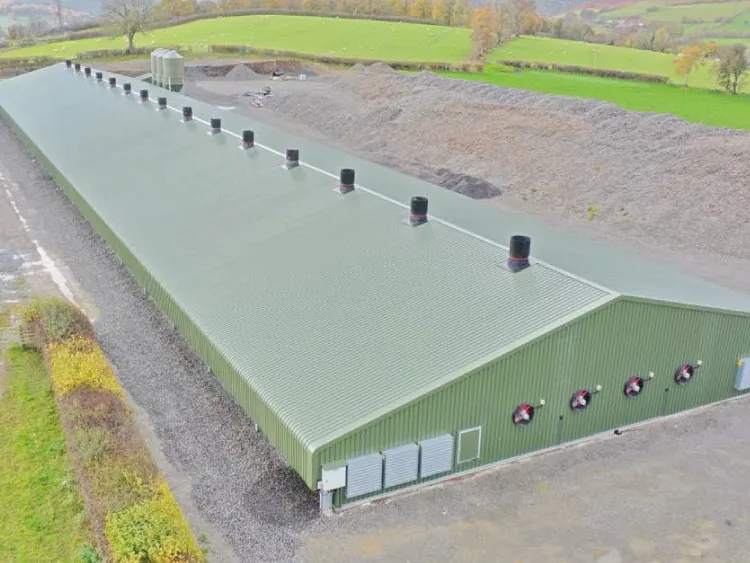 Large green industrial or agricultural building with multiple roof vents and wall-mounted fans in a rural area.