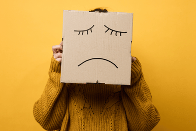 A person wearing a mustard-colored sweater holds a cardboard box with a sad face drawn on it in front of their face against a yellow background.