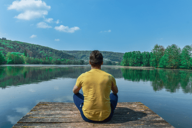 A person sits in a meditative pose on a wooden dock overlooking a calm lake with lush green hills in the background.