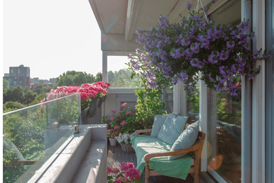 A balcony with a wooden bench, light blue cushions, and a green throw blanket, surrounded by potted pink and purple flowers under a bright sky.