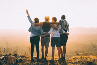 Four friends stand with arms around each other on a hilltop at sunset, with one person raising an arm in the air.