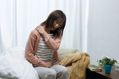 A person sitting on a bed with their hand on their shoulder, appearing to be in pain.