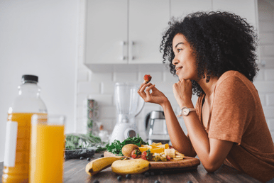 A person holds a strawberry while looking at a platter of sliced fruit including kiwi, oranges, and bananas, with a bottle and glass of orange juice nearby.