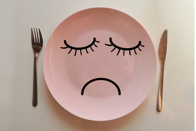 A pink plate with a sad face drawn on it, placed between a fork and a knife.