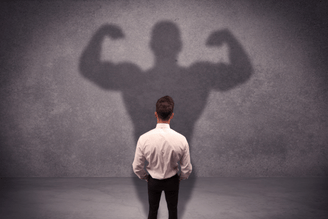 A person in a white shirt and black pants stands with their back to the viewer, casting a large shadow on a concrete wall that depicts a muscular figure flexing its biceps.