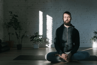 A man with a beard meditates cross-legged on a yoga mat in a sunlit room with plants.