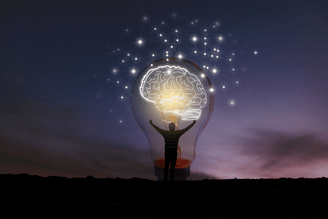 Silhouette of a person with arms raised inside a lightbulb containing a glowing brain illustration and connected network of lights against a twilight sky.