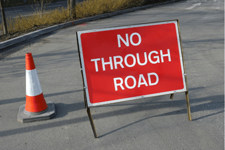 A red and white traffic cone stands next to a red "NO THROUGH ROAD" sign on an asphalt surface.