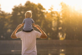 A person carries a child on their shoulders near a body of water with trees in the background during golden hour.