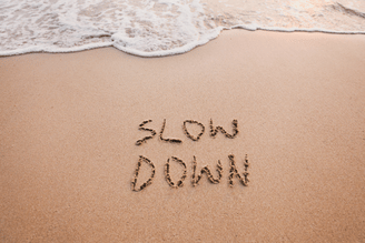 The words "SLOW DOWN" are written in the sand on a beach, with gentle waves washing ashore.