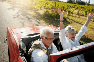 A smiling couple in a red convertible drives down a road lined with vineyards, with the passenger raising their arms in excitement.