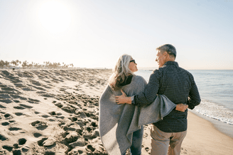 A couple walks arm-in-arm along a sandy beach next to the ocean under a bright sky.